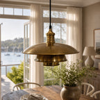 The Mercantile Brass Pendant in antique, softly aged brass hangs above a wooden dining table in a sunlit kitchen.