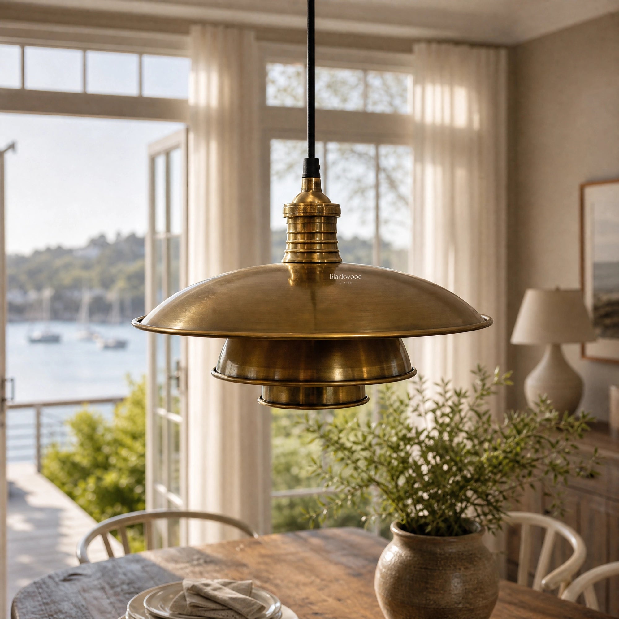 The Mercantile Brass Pendant in antique, softly aged brass hangs above a wooden dining table in a sunlit kitchen.