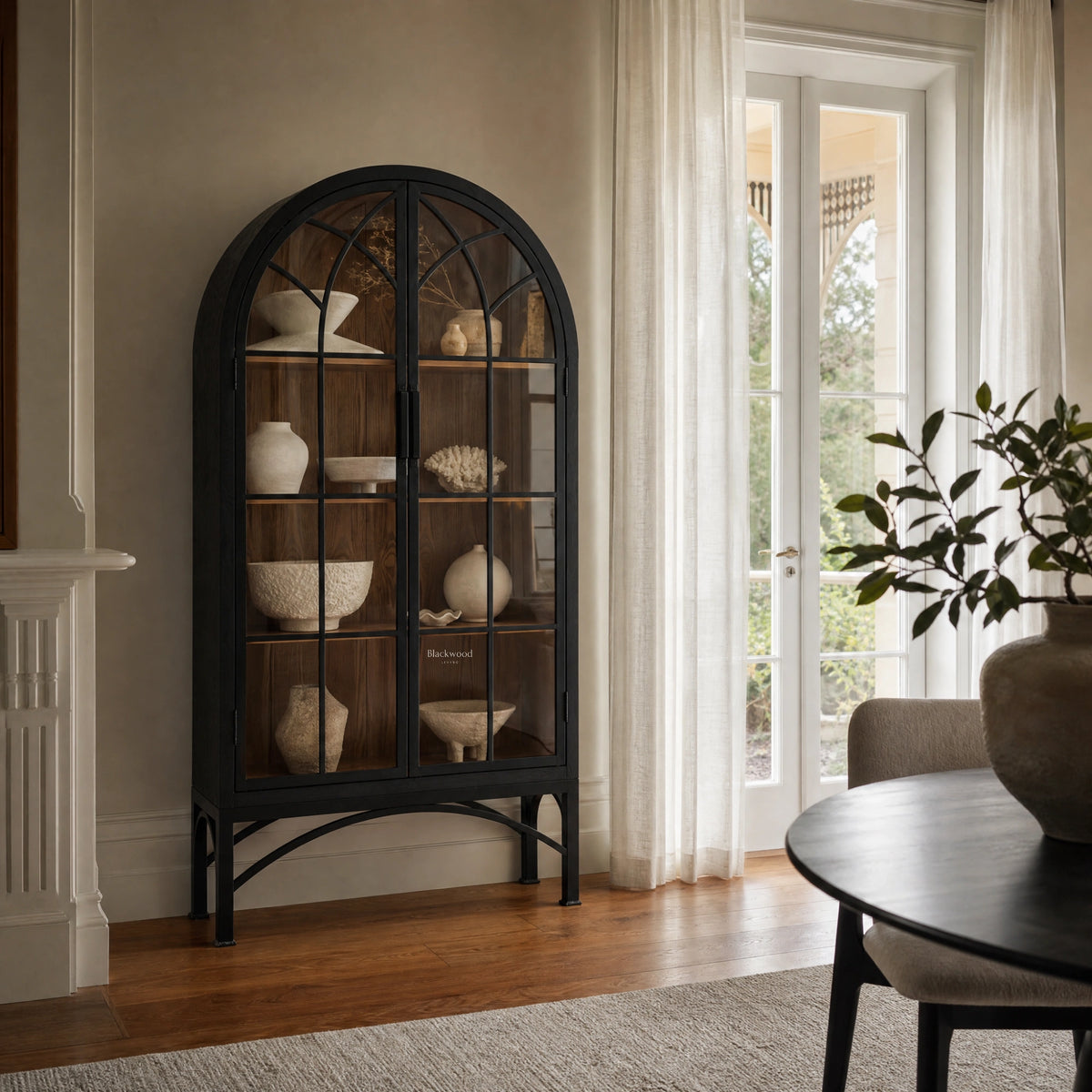 Tall black arched display cabinet with glazed doors and metal base holding neutral ceramic vessels.