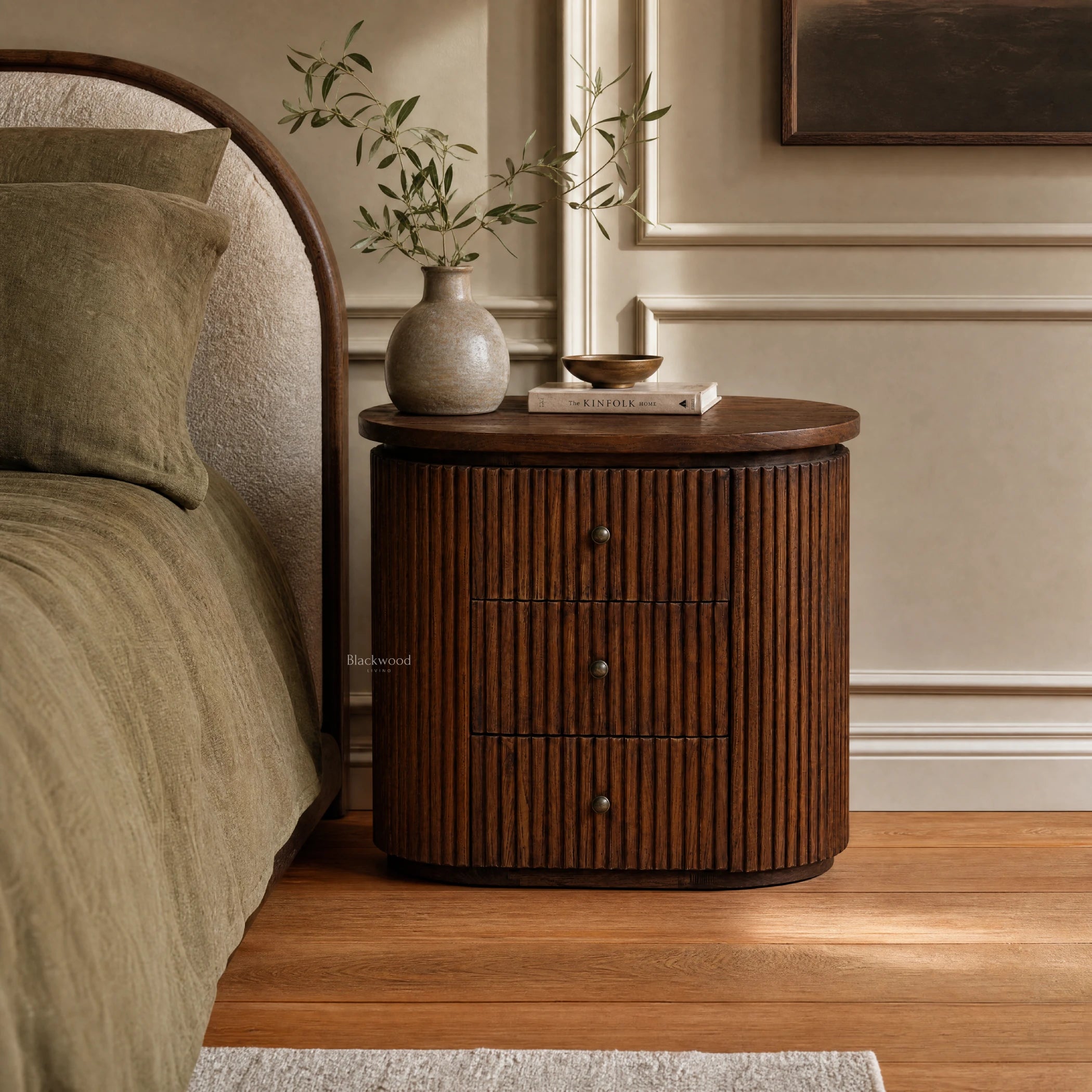 Reed Bedside Table in old elm with a rounded silhouette and deeply fluted timber detailing beside a bed.