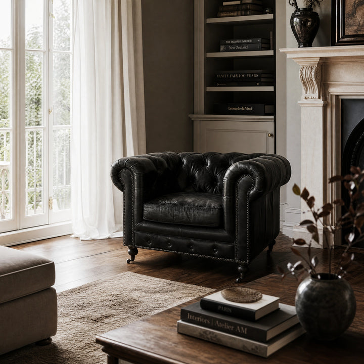 Ashcroft Armchair in estate black leather with Chesterfield tufting in a living room by bookshelf and fireplace.