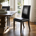Manor Dining Chair in black leather with brass-stud trim, solid wood legs at a sunlit dining table.