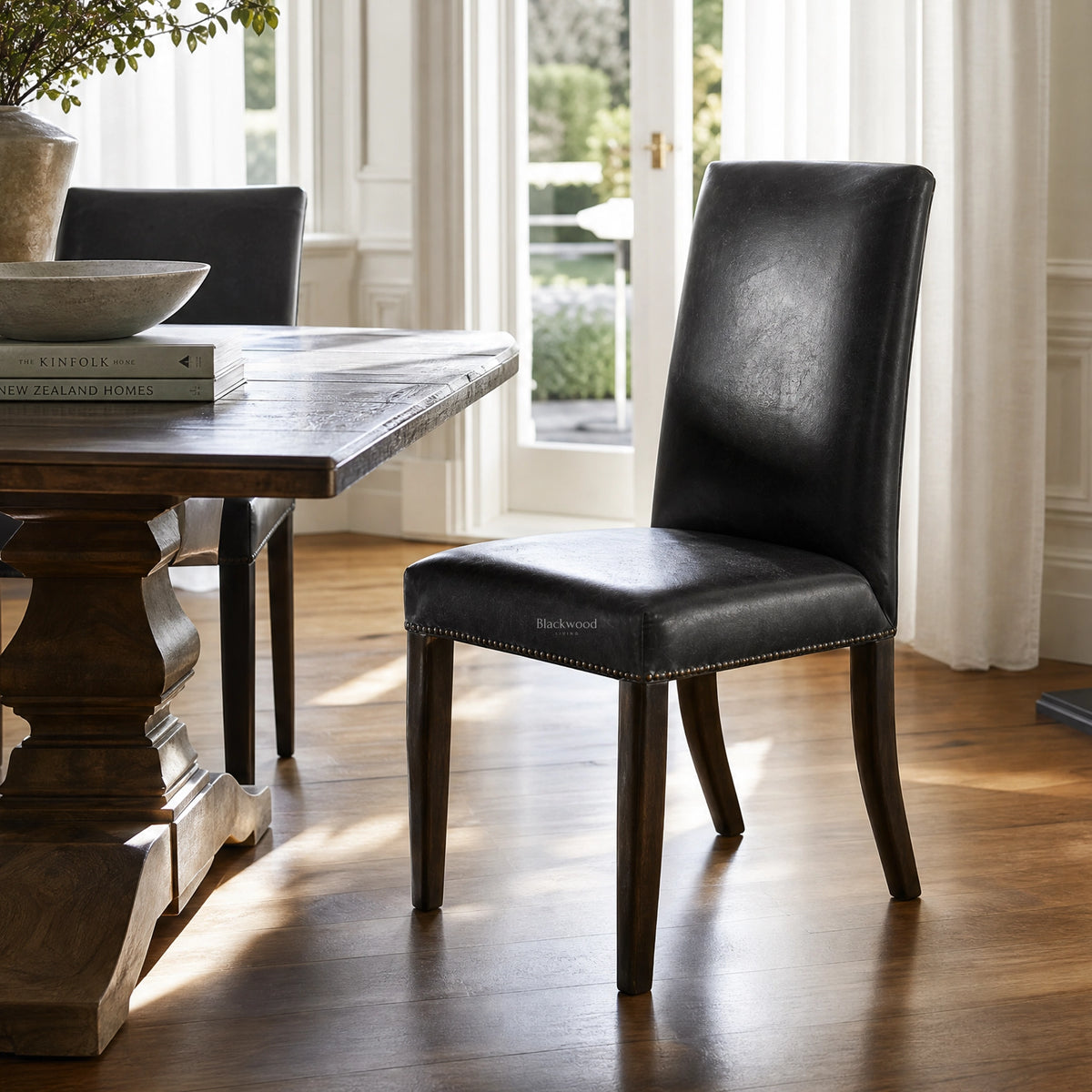 Manor Dining Chair in black leather with brass-stud trim, solid wood legs at a sunlit dining table.