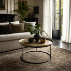Round antique brass coffee table with raised tray edge and slender black iron base in a living room setting.
