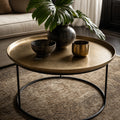 Round antique brass coffee table with slender black iron legs, styled with dark ceramic decor and a monstera leaf on a textured rug.