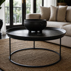 Round black coffee table with raised tray top and slender iron frame, styled with a decorative bowl in a living room.